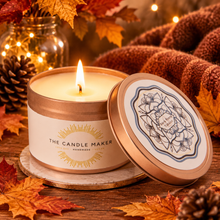 Load image into Gallery viewer, A lit candle in a round tin with the lid leaning beside it on a wooden surface. The candle label reads 'THE CANDLE MAKER' and 'HANDMADE.