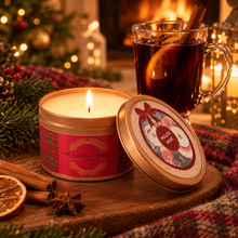 Load image into Gallery viewer, A lit holiday-themed candle in a round tin labeled 'The Candle Maker' and 'Spilled Wine' scent, placed on a wooden surface beside a glass of hot mulled wine, with cinnamon sticks, star anise, pine branches, and warm festive lights in the background.