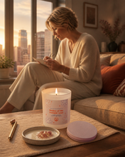 Load image into Gallery viewer, A woman sits on a couch in a warmly lit living room, writing in a notebook beside a lit candle on a table. The candle jar has a white label with pink floral details and the text 'Mother of the freakin' you' and 'sophia'.
